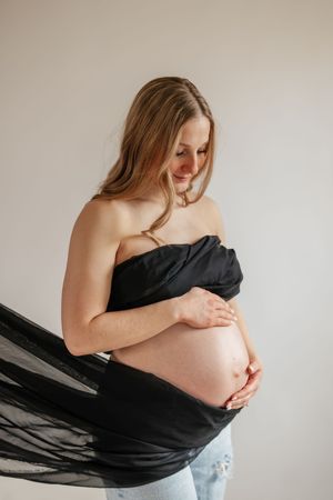 Pregnant woman in black wrap top and jeans cradling her bare baby bump, smiling downward against a light background.
