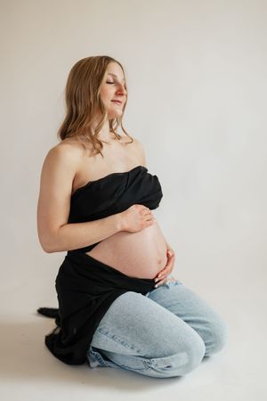 Pregnant woman in black wrap top and jeans kneeling, cradling her bare baby bump against white background.