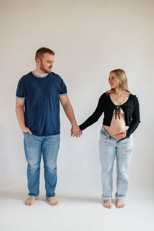 Pregnant couple holding hands barefoot against white background, man in navy shirt and jeans, woman in black top showing baby bump.