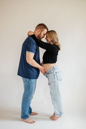 A couple in jeans share a tender forehead-to-forehead moment, hands intertwined, against a white background.