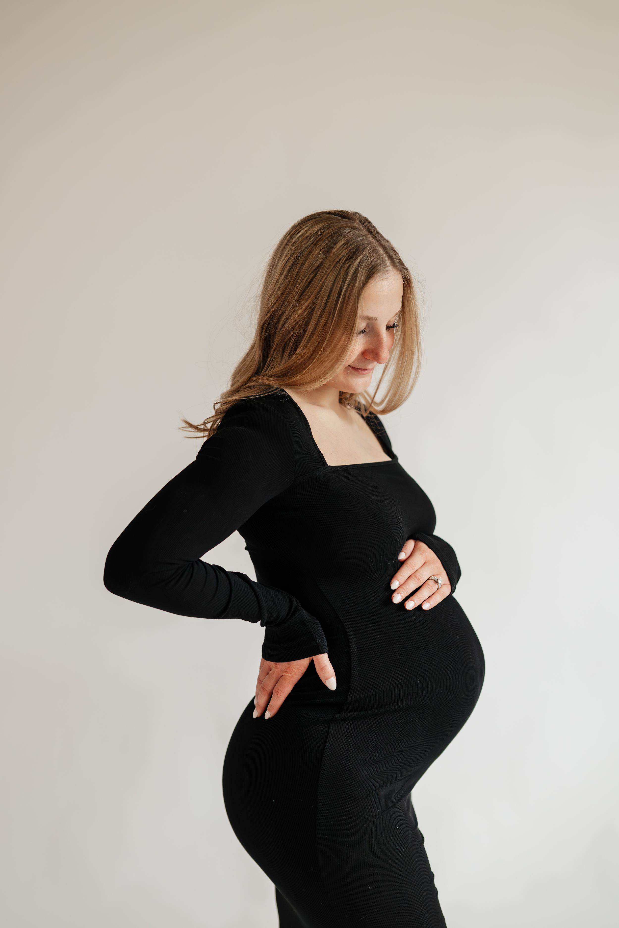 Pregnant woman in black long-sleeve dress cradling baby bump, smiling downward against white background.