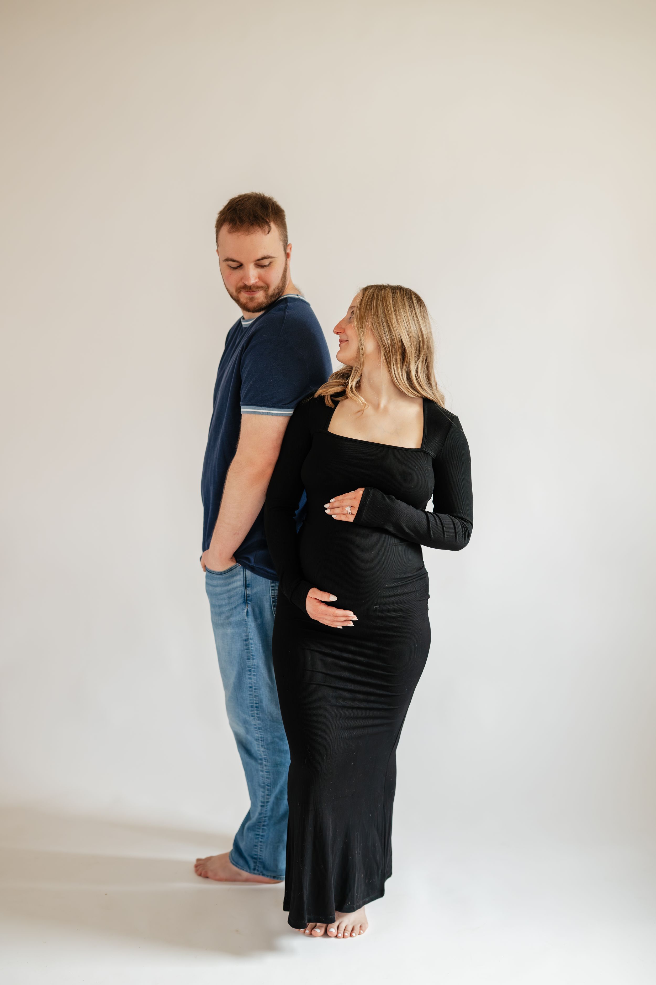 Pregnant woman in black dress stands back-to-back with man in blue shirt and jeans, both smiling in studio.