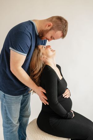 A pregnant woman in black sits smiling as her partner in a blue shirt tenderly leans in close in a studio setting.