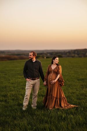 Pregnant couple holding hands in a green field at golden hour sunset, woman in elegant brown maternity gown.
