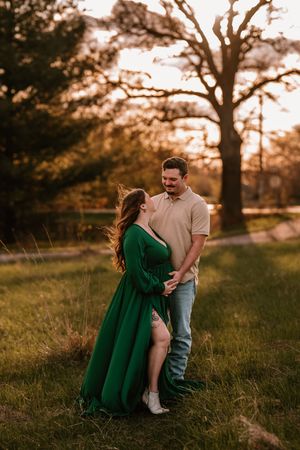 Couple embracing at golden hour in a park, woman in elegant green dress, man in beige shirt and jeans, trees in background.