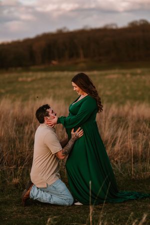 A man kneels before a pregnant woman in a green dress during a golden hour maternity photoshoot in a scenic field.