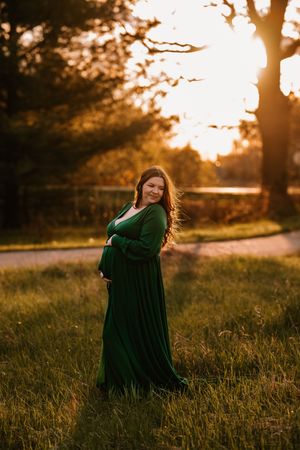 Pregnant woman in emerald green gown standing in sunlit park at golden hour, cradling baby bump with flowing red hair.