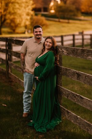 Couple poses by wooden fence at golden hour, woman in emerald green maternity gown cradling baby bump.