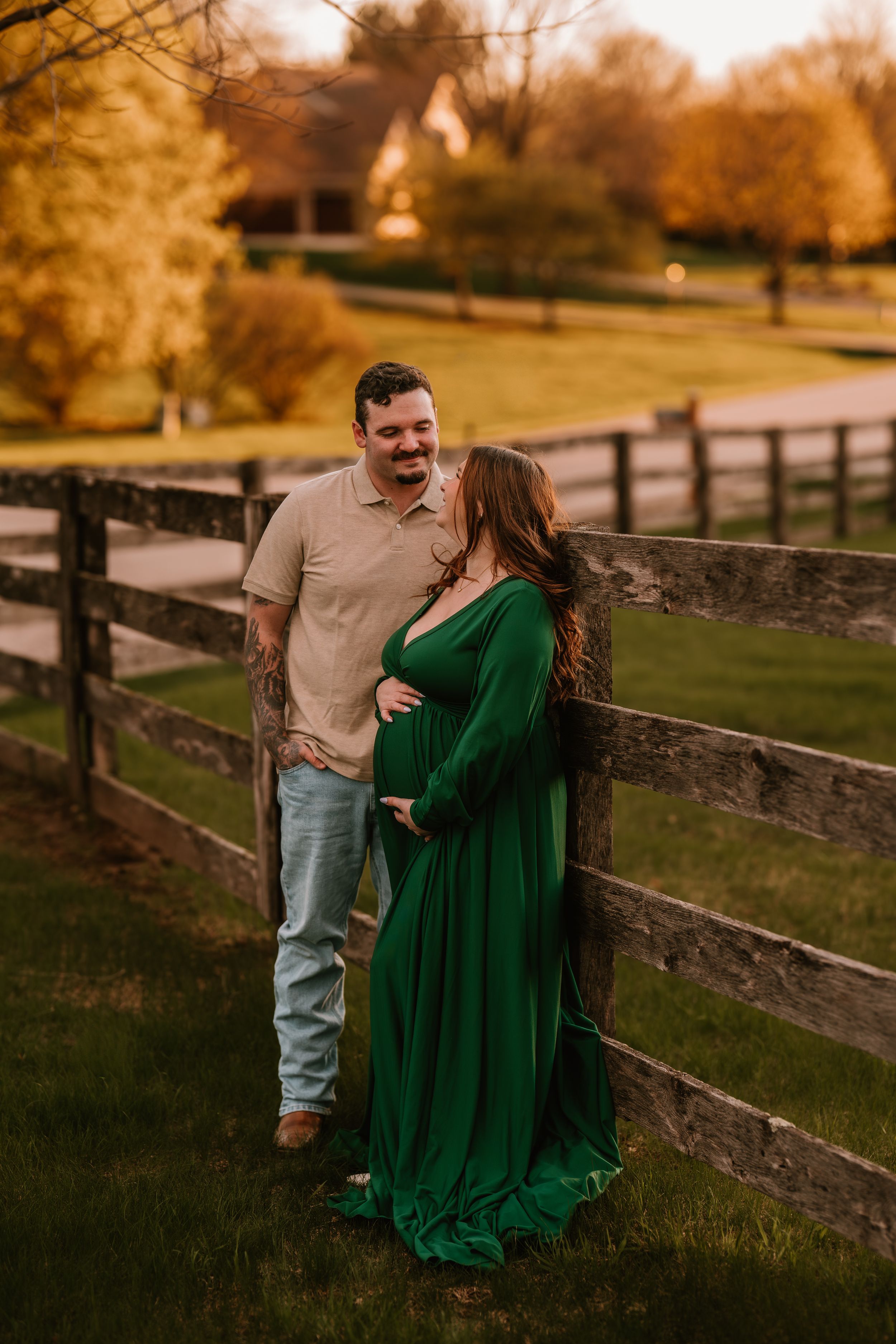 Couple laughing together at golden hour by a wooden fence, woman in elegant green maternity gown on a lush farm.