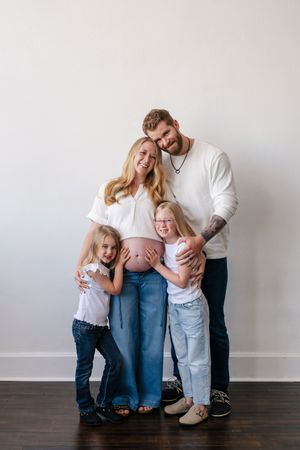 Pregnant woman with exposed belly poses with husband and two young daughters in matching white tops and jeans for maternity photo.