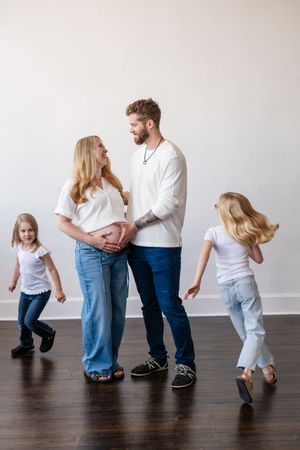 Pregnant woman and bearded man in white shirts and jeans smile at each other while two young girls play around them.