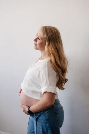 Pregnant woman in white top and jeans cradling her baby bump, smiling serenely in a bright, minimalist setting.