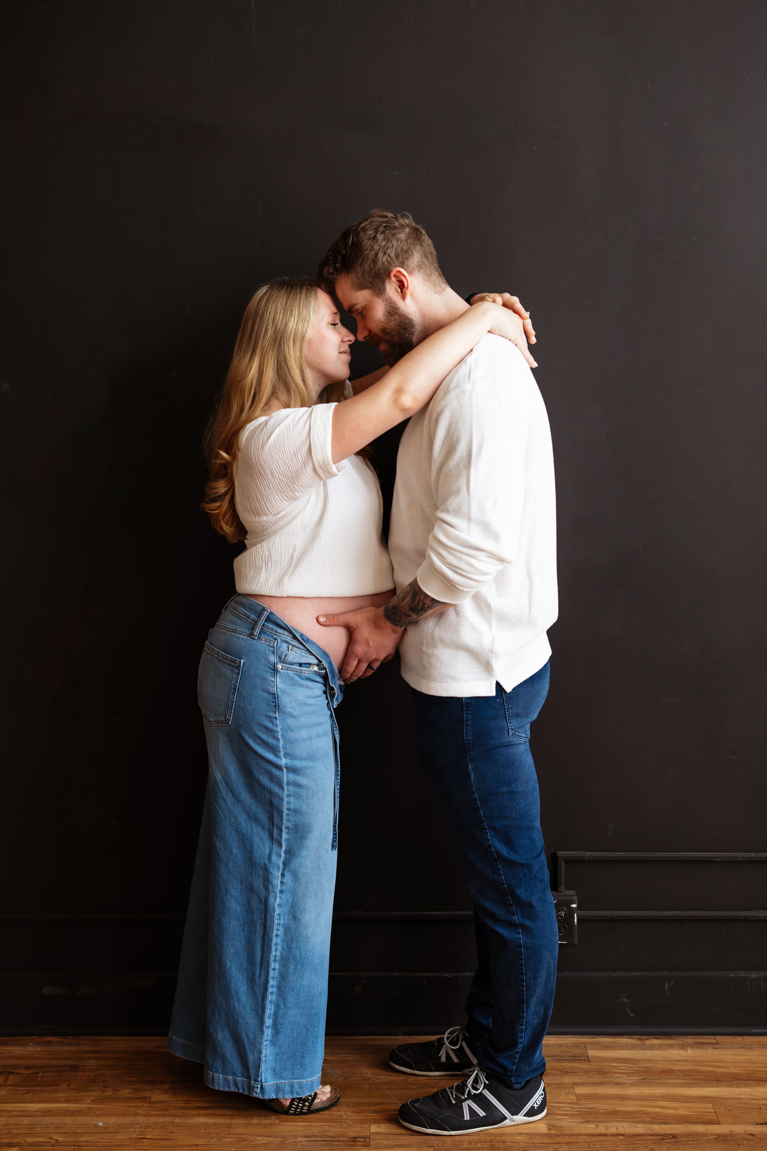 A couple in casual denim and white tops share an intimate embrace against a dark wall on a wooden floor.