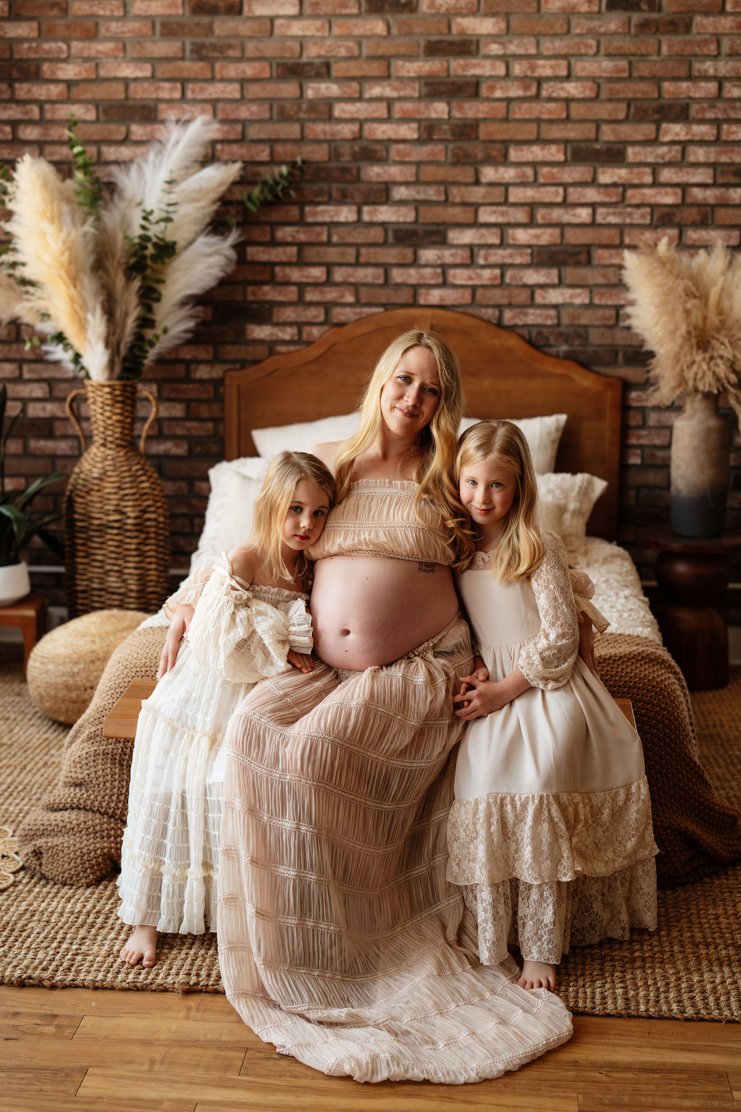 Pregnant blonde woman in beige lace gown sits on bed with two young daughters in white dresses, brick wall backdrop.