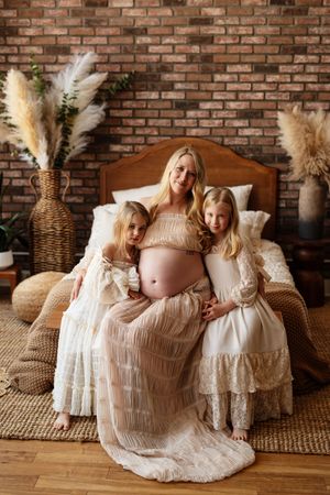 Pregnant blonde woman in beige lace gown sits on bed with two young daughters in white dresses, brick wall backdrop.