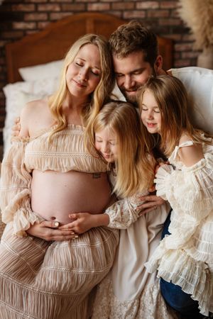 Pregnant woman in lace dress with husband and two daughters tenderly embracing baby bump in cozy bedroom setting.