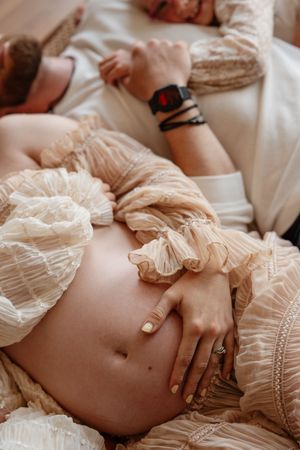 Pregnant woman in a beige ruffled dress lying down, partner beside her with hand on baby bump.
