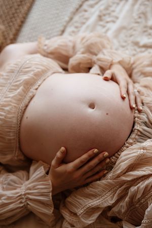 Close-up of pregnant woman cradling bare baby bump in elegant cream lace dress during maternity photoshoot.
