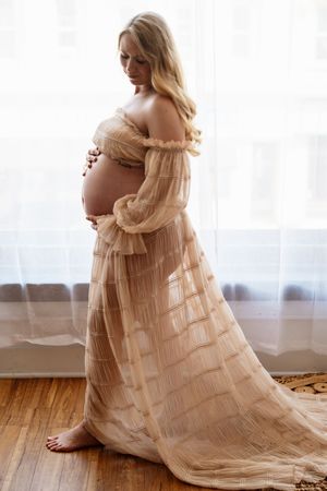 Pregnant woman in elegant beige off-shoulder tulle gown standing by window, cradling baby bump in soft natural light.