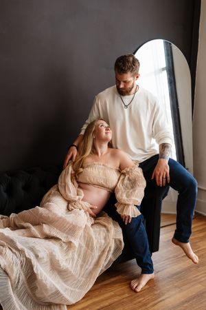 Pregnant woman in beige dress reclines against man in white sweater during maternity photoshoot in dark studio.