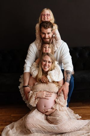 Pregnant woman in beige dress sits on floor with tattooed husband and two young daughters stacked behind her.