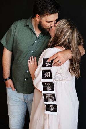 Couple kissing during pregnancy announcement photo shoot, woman in white dress holding ultrasound sonogram strip, black background.