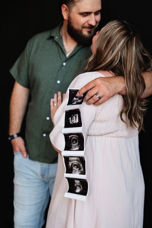 Pregnant couple embracing while holding ultrasound photos against baby bump during maternity photo shoot.