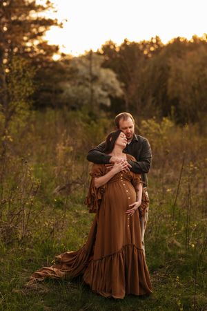 Couple embracing at golden hour in a field, pregnant woman in brown dress with man holding her belly.