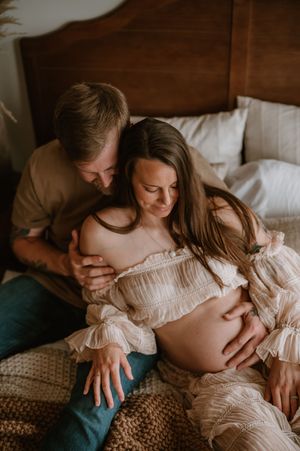 Couple sharing a tender maternity photo session on a bed, with man embracing pregnant woman in a ruffled cream off-shoulder dress.