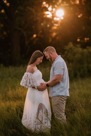 Couple sharing a tender moment in a sunlit field at golden hour, woman in white floral dress cradling baby bump.