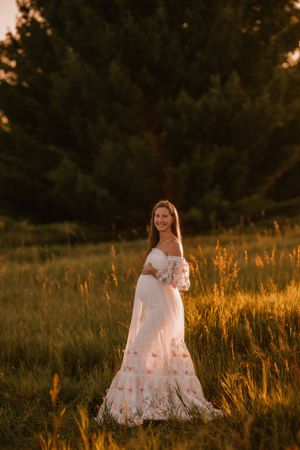 Pregnant woman in white off-shoulder floral gown standing in golden sunset field with lush green trees backdrop.