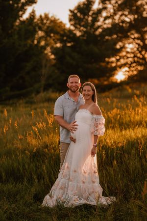 Couple poses in golden sunset field, woman in white floral off-shoulder maternity gown, man in blue shirt embracing her bump.