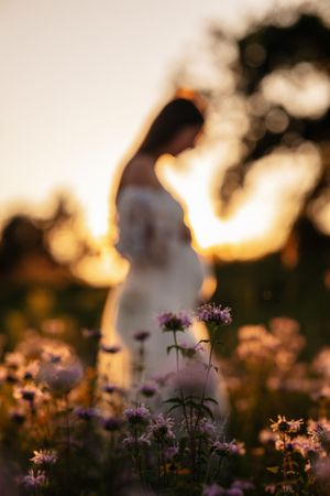 Pregnant woman in white dress standing in a wildflower field at golden sunset, soft bokeh purple flowers in foreground.