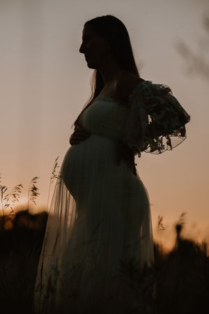 Silhouette of a pregnant woman in a white dress holding flowers at golden hour in a field.