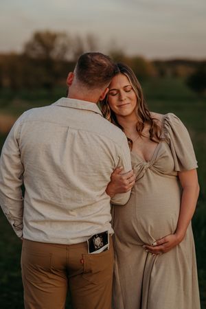 Couple sharing a tender maternity moment outdoors at golden hour, man with sonogram in pocket, pregnant woman in beige dress.