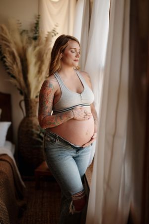 Pregnant woman with tattoos cradling baby bump, wearing gray bralette and jeans, standing by sheer curtains.