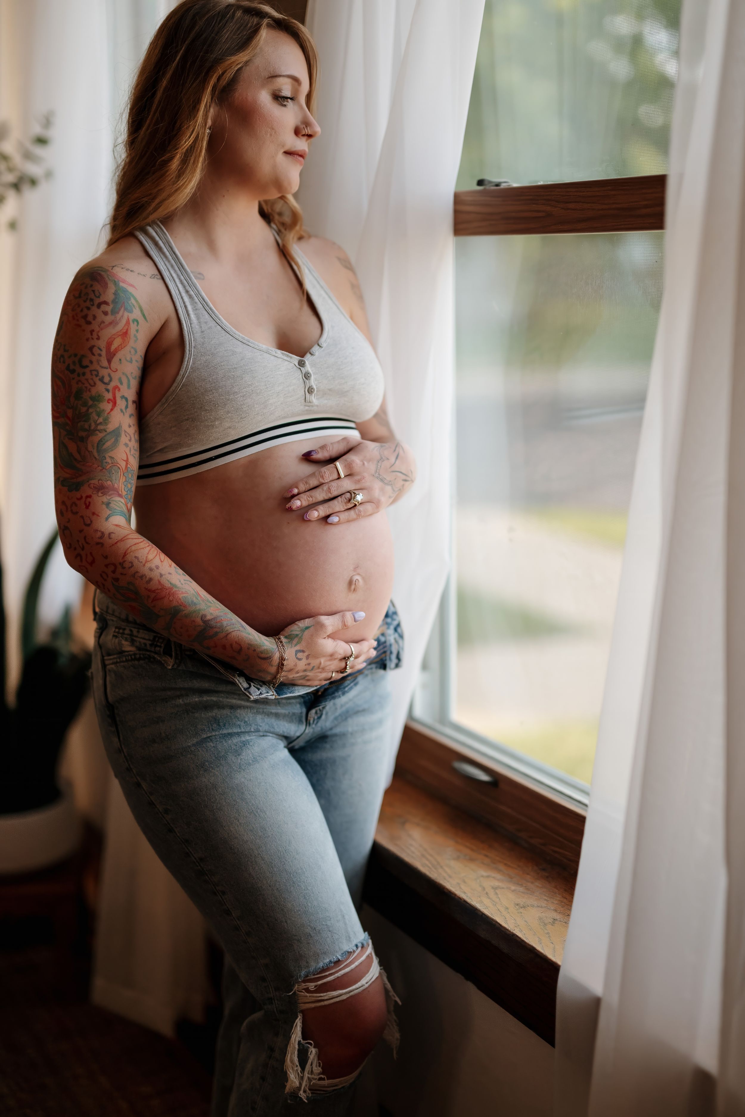 Tattooed pregnant woman in gray crop top and jeans cradling baby bump by a sunlit window.
