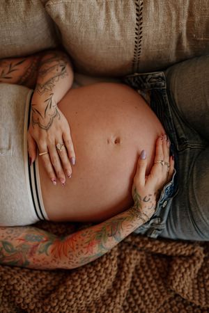 Pregnant woman with tattoos cradling her bare baby bump, wearing jeans and white underwear, resting on a couch.