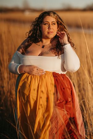 Woman in white off-shoulder top and colorful maxi skirt poses in golden field at sunset.