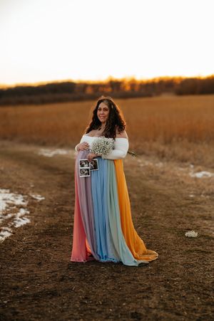 Pregnant woman in rainbow maternity gown holding ultrasound photos and flowers in a golden field at sunset.