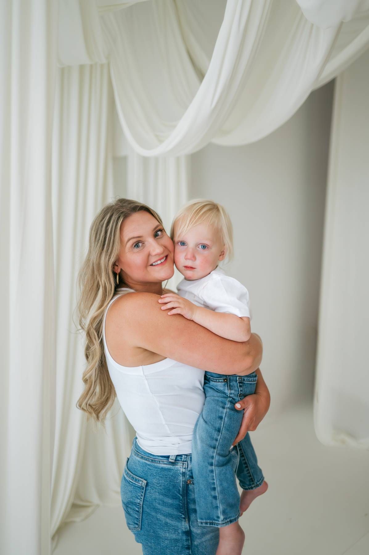 Smiling blonde mother holding adorable toddler in white studio with flowing white fabric backdrop in North Carolina