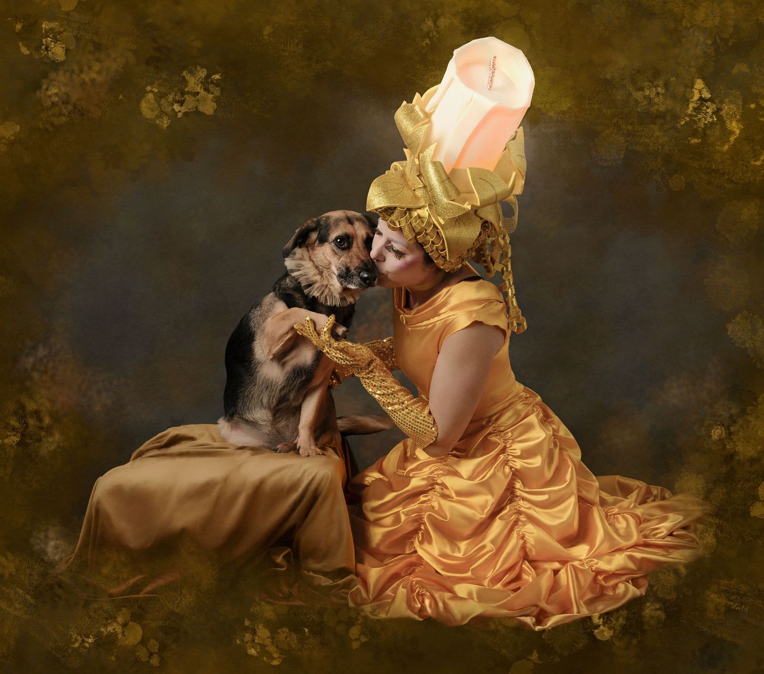 A woman in a golden gown and floral headpiece shares a tender moment kissing a small dog in a vintage portrait.