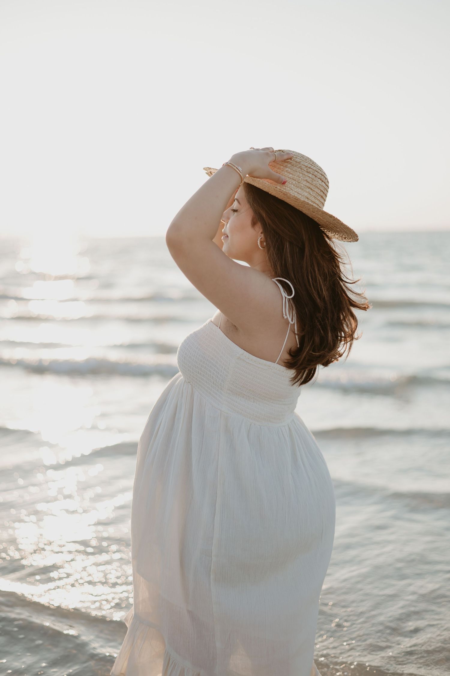 Woman in white dress holding straw hat at sunny beach with ocean waves in background.