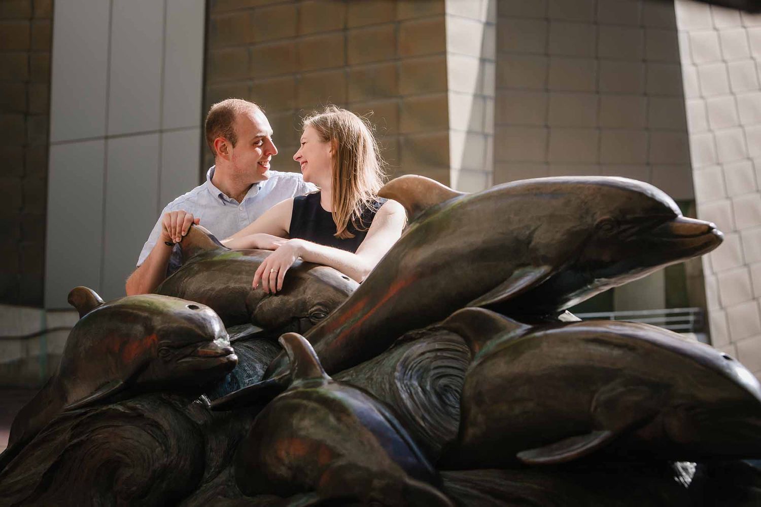 Engaged couple laughing together among the bronze dolphin statues outside the New England Aquarium in Boston