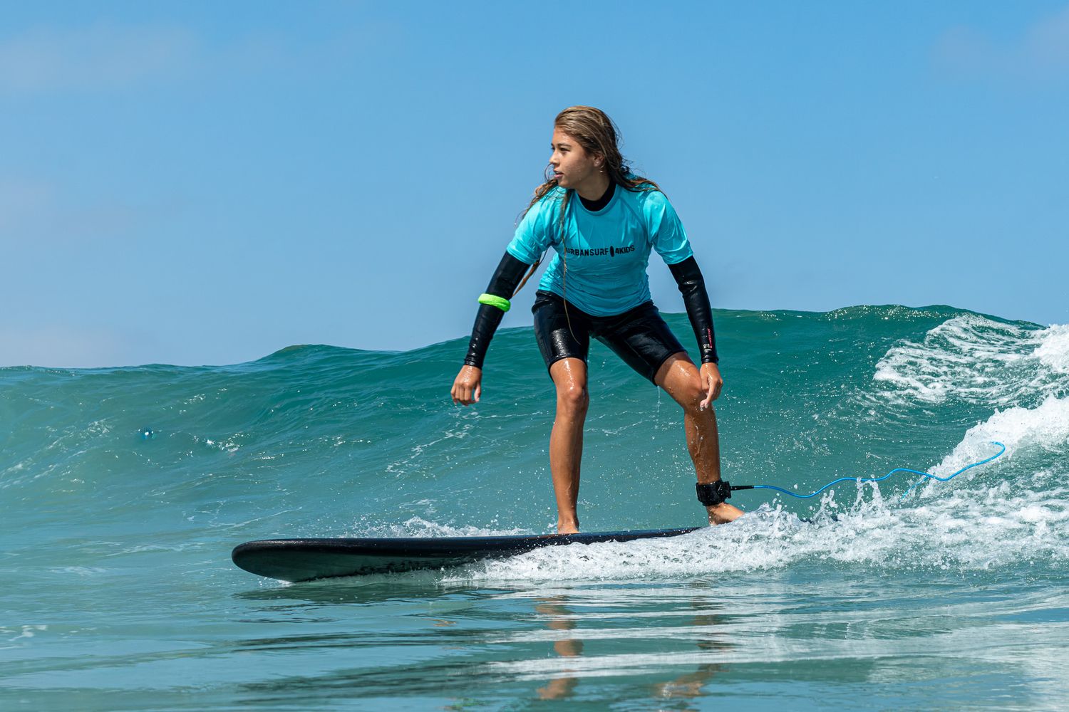 Young surfer riding a wave on a black surfboard wearing a teal wetsuit on a sunny day.
