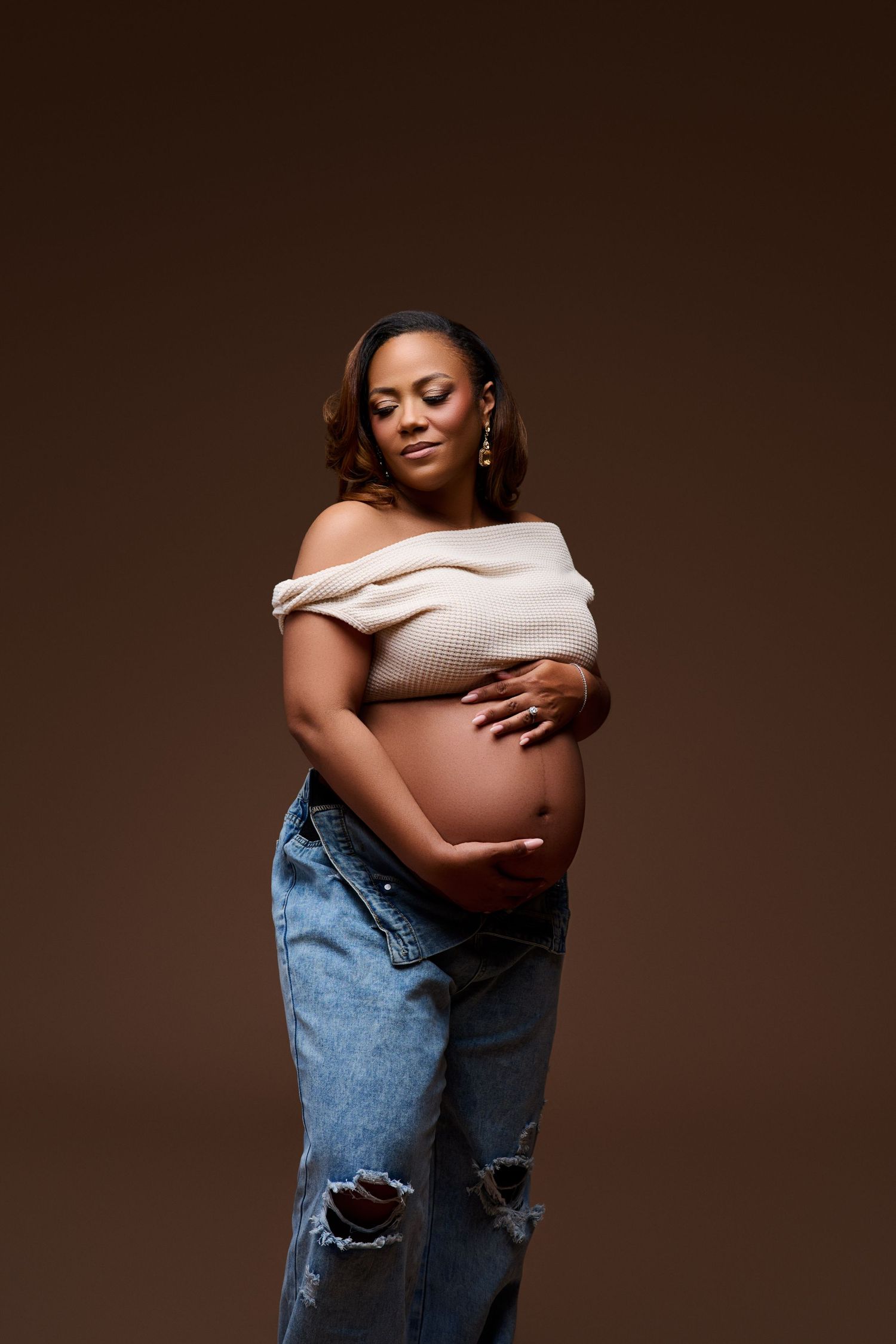 Pregnant Black woman in off-shoulder top and ripped jeans cradling her baby bump against a brown studio background. NJ Maternity Photography