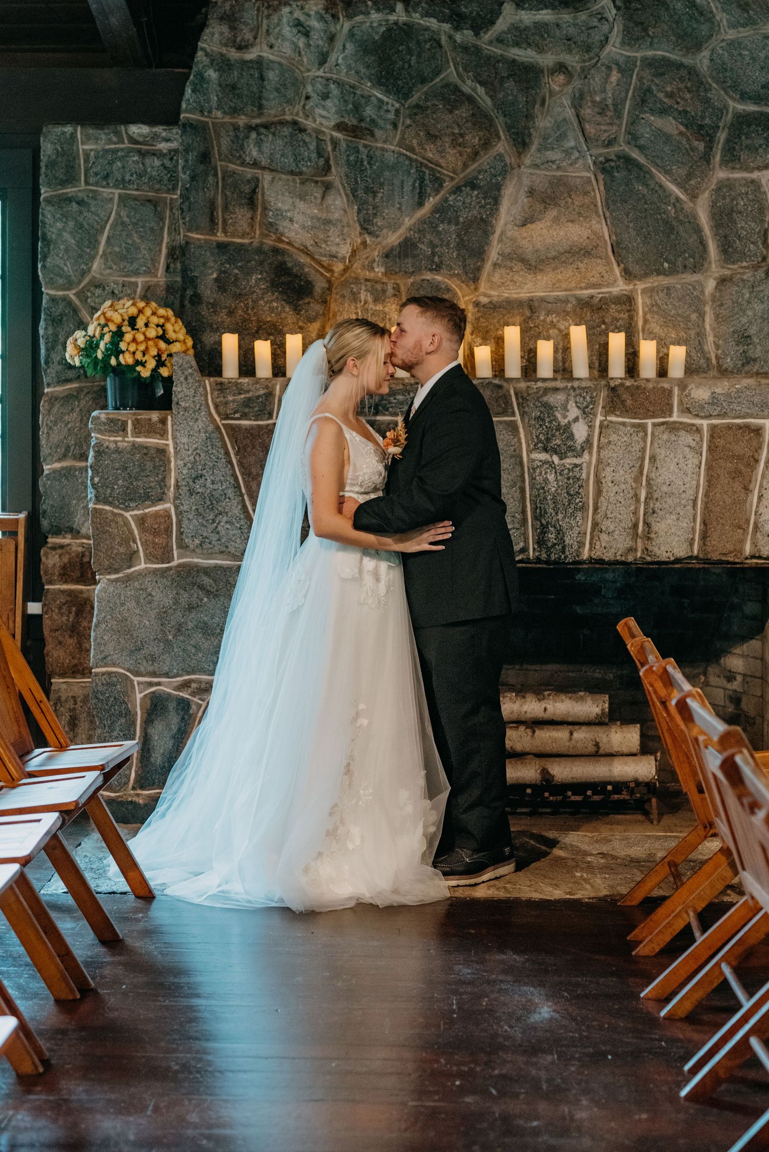 Bride and groom share first kiss at rustic stone fireplace wedding ceremony with candles and wooden chairs.