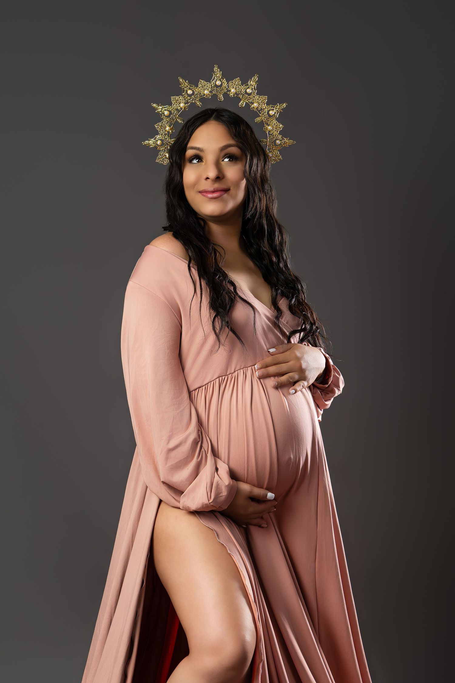 Elegant pregnant woman in pink flowing gown wearing gold crown poses gracefully in studio portrait.