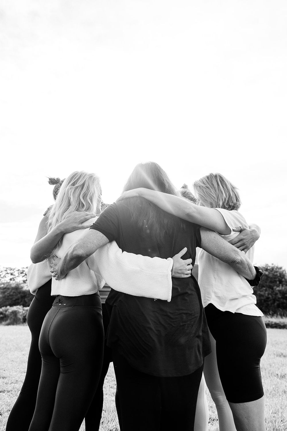 Black and white photo of a group of women hugging in a circle outdoors, viewed from behind.
