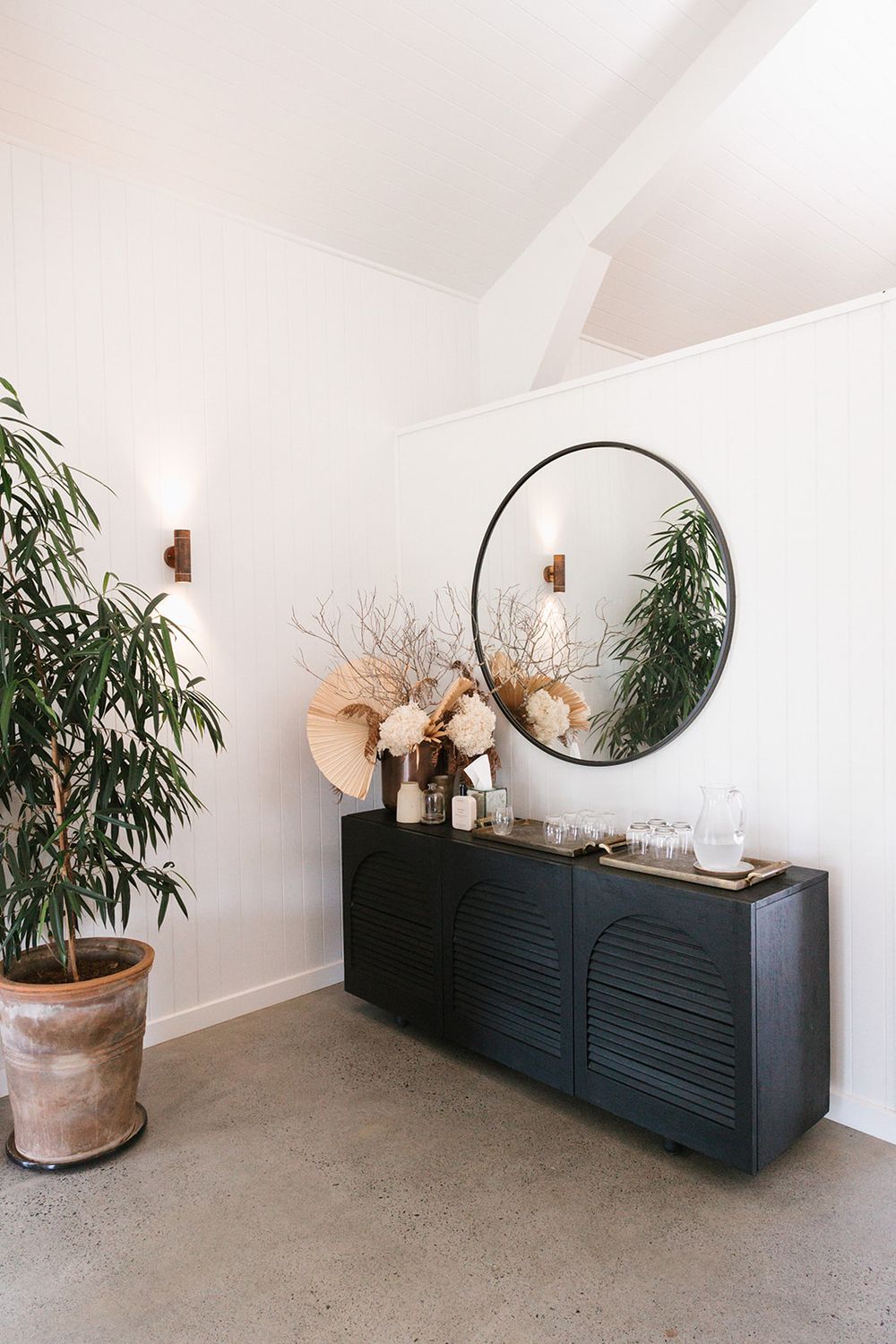 Modern entryway with black sideboard, round mirror, wall sconces, dried floral arrangement, and potted green tree on concrete floor.